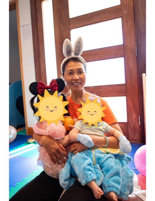 A smiling Little Star caregiver holding two children, whose faces are covered with stickers