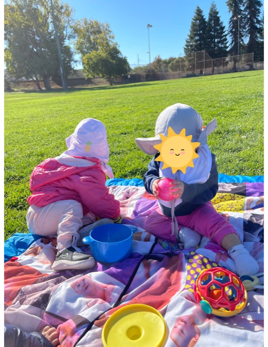 Two children playing on a picnic blanket in a grassy park; face stickers are present
