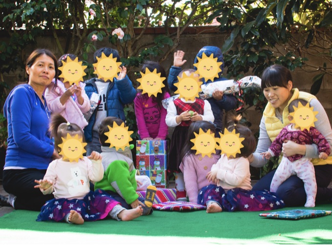 Caregivers sitting with children in the shaded backyard; the children’s faces have stickers