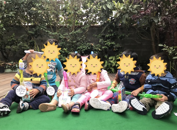 Children sitting together outdoors beneath leafy trees; their faces have stickers